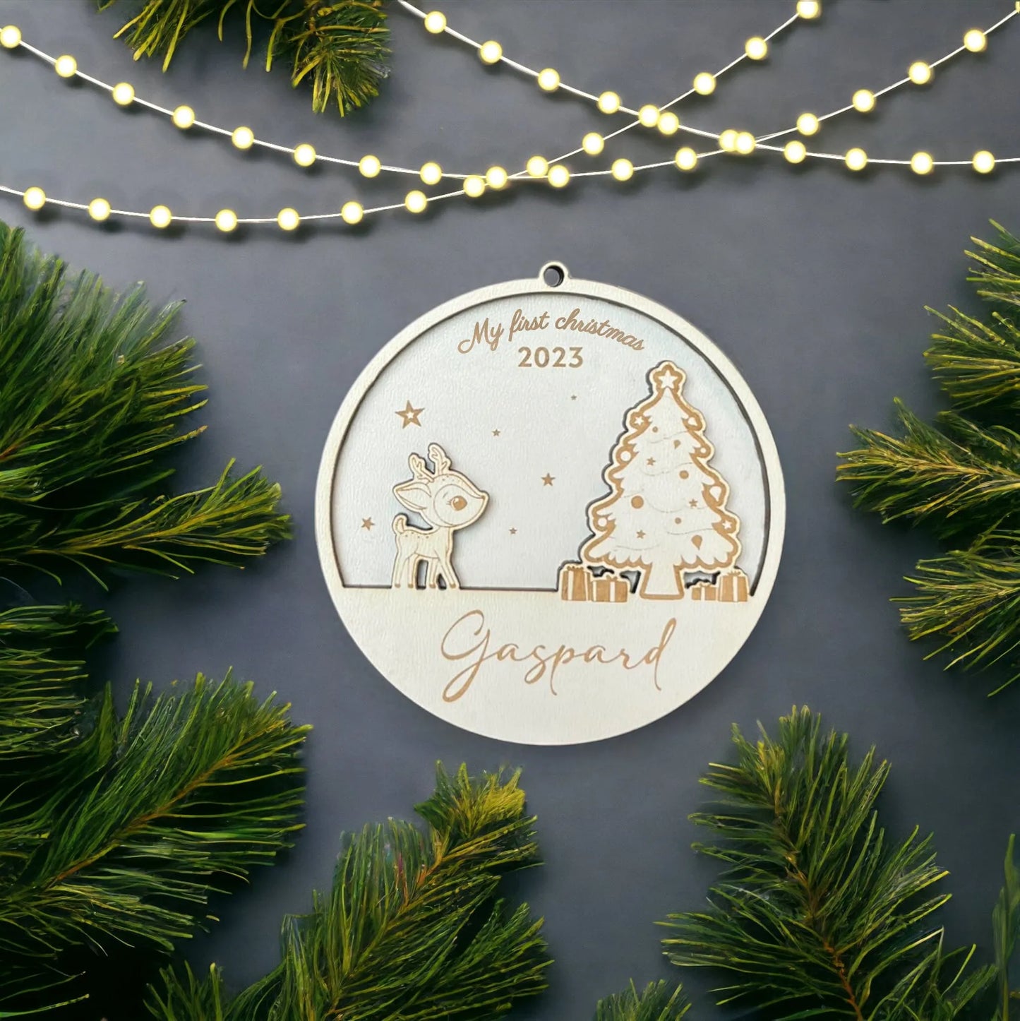 Decoration de Noel circulaire en bois, gravée avec "My First Christmas 2023" en haut et le prénom "Gaspard" en bas. L'ornement présente un petit renne et un sapin de Noel décoré, avec des cadeaux à la base du sapin. La décoration est placée sur un fond gris, entourée de branches de sapin et illuminée par une guirlande lumineuse, créant une ambiance festive et chaleureuse