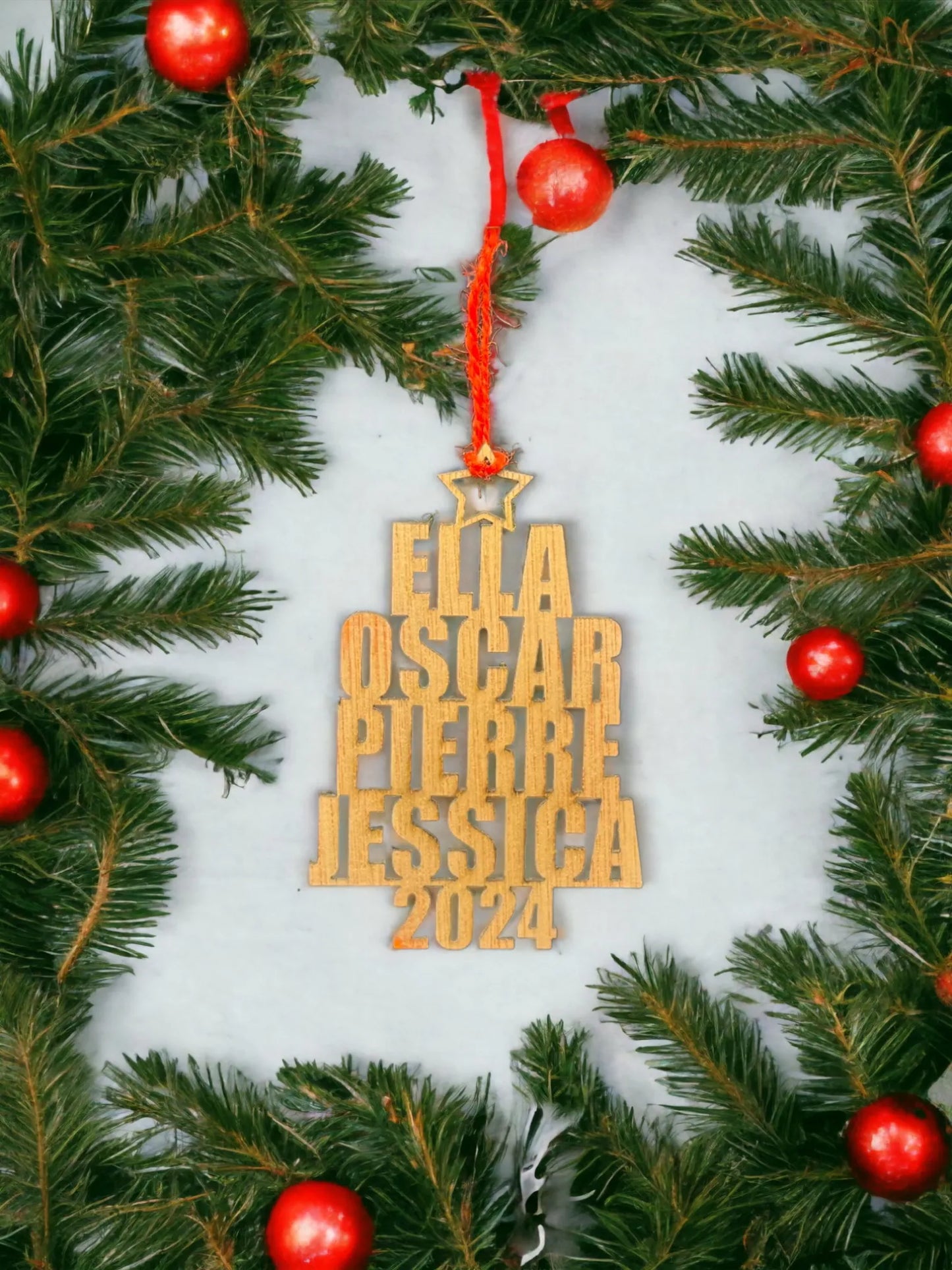 Décoration en bois suspendue, gravée avec les prénoms "Ella", "Oscar", "Pierre", "Jessica", et l'année "2024". L'ornement est entouré de branches de sapin avec des boules rouges, offrant une touche festive parfaite pour une décoration de Noël. Le contraste du bois clair et des ornements rouges ajoute un charme rustique et personnalisé à l'ensemble.