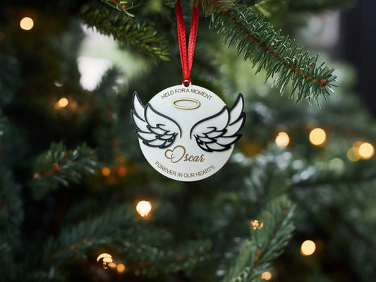 décoration de Noël en forme de médaillon suspendue à une branche de sapin, ornée d'un motif d'ailes d'ange avec un halo doré au-dessus. Le nom "Oscar" est gravé en bas, accompagné de la phrase "Held for a moment, forever in our hearts"