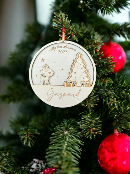Une decoration de Noel en bois suspendue à un sapin, gravée avec le texte "My First Christmas" en haut et le prénom "Gaspard" en bas. Le design inclut un petit renne et un sapin décoré, entourés de cadeaux et d'étoiles. La décoration est suspendue à un ruban rouge, ajoutant une touche festive et personnalisée pour célébrer le premier Noel de l'enfant.