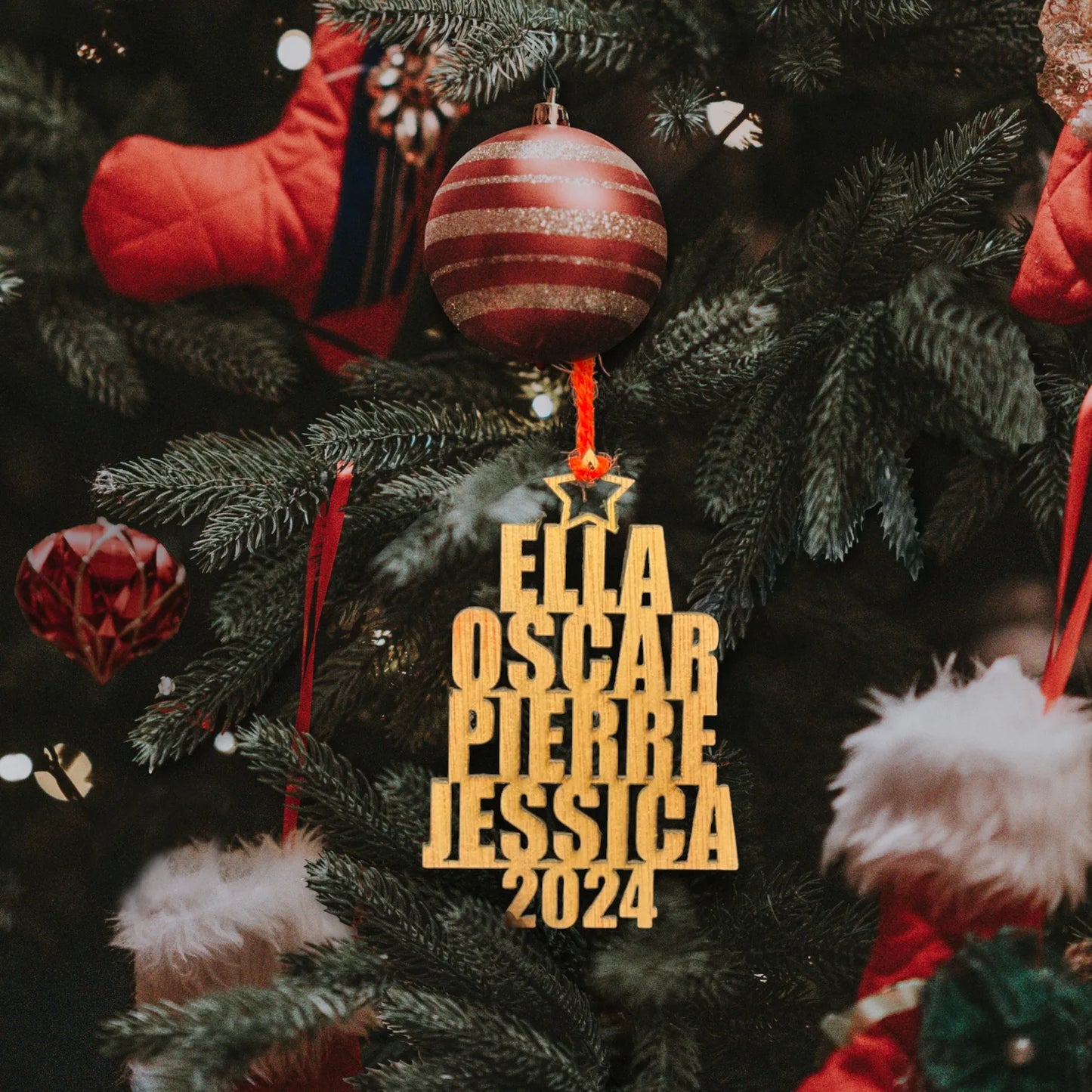 Décoration en bois suspendue à un sapin de Noël, affichant les prénoms "Ella", "Oscar", "Pierre", et "Jessica" gravés verticalement avec l'année "2024" en bas. La décoration est accrochée avec un ruban rouge, tandis que des boules rouges et d'autres ornements décoratifs de Noël, comme des bas, complètent l'arrière-plan festif.