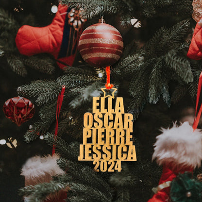 Décoration en bois suspendue à un sapin de Noël, affichant les prénoms "Ella", "Oscar", "Pierre", et "Jessica" gravés verticalement avec l'année "2024" en bas. La décoration est accrochée avec un ruban rouge, tandis que des boules rouges et d'autres ornements décoratifs de Noël, comme des bas, complètent l'arrière-plan festif.