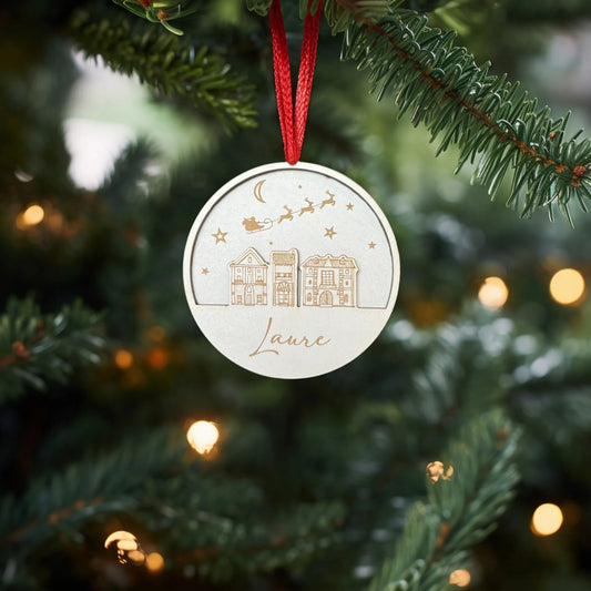 Decoration de Noel en bois suspendue a un sapin, gravée avec un design représentant des maisons sous un ciel étoilé, avec un traineau tiré par des rennes volant dans les airs. Le prénom "Laure" est inscrit en bas, gravé de manière élégante. La decoration est accrochée à un ruban rouge, ajoutant une touche personnalisée et festive à l'arbre de Noel, entourée de lumières scintillantes.