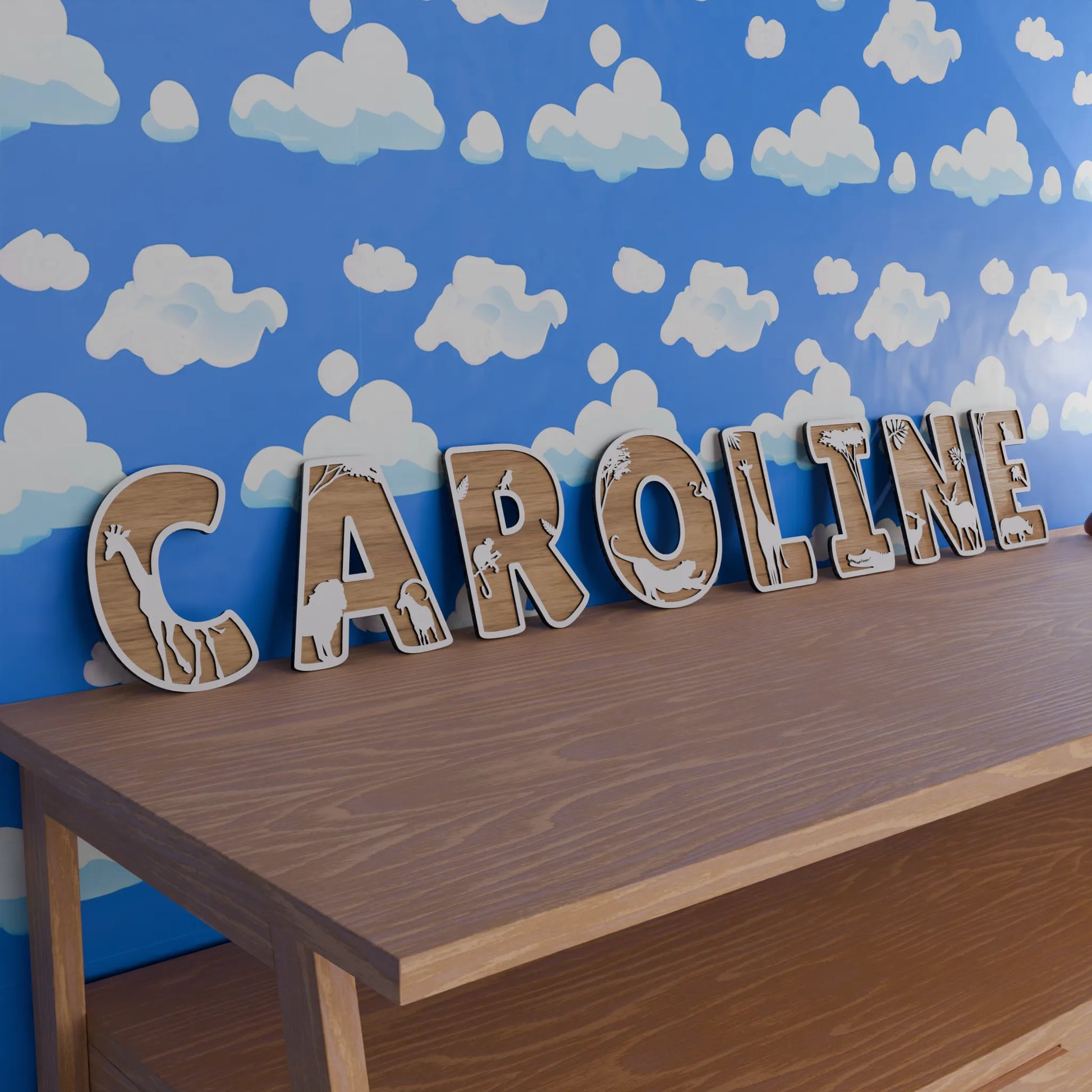 lettres en bois découpées formant le prénom "Caroline", chaque lettre étant décorée d'un motif d'animaux sauvages dans un style safari, avec des girafes, éléphants, et autres créatures. Les lettres sont posées sur une étagère en bois devant un mur peint de nuages blancs sur un fond bleu ciel, créant une ambiance de chambre d'enfant avec un thème naturel et aventureux.